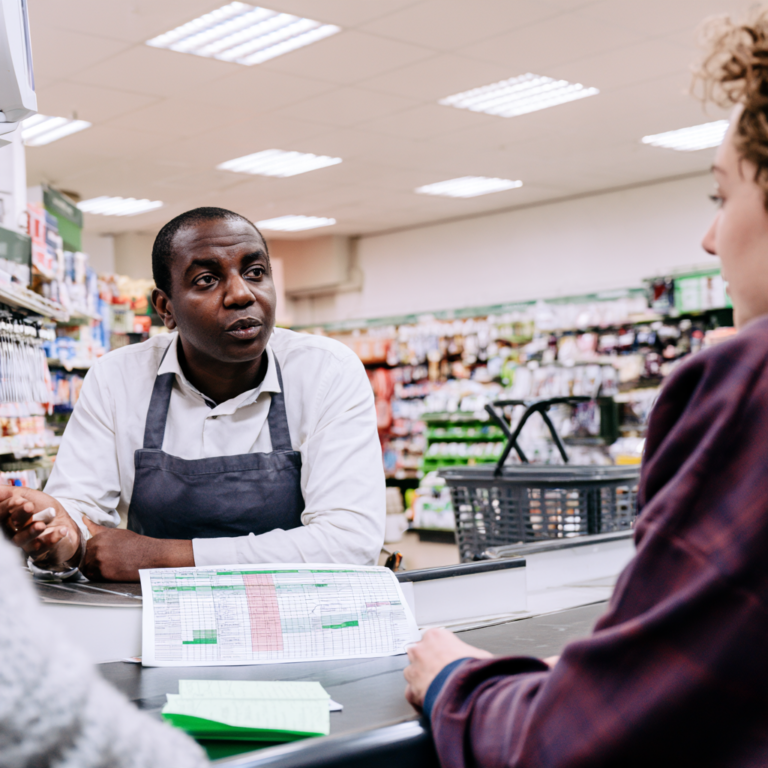 Butikschef och sökande samtalar vid kassadisken i en butik, med veckoschema på en pärm framför sig.